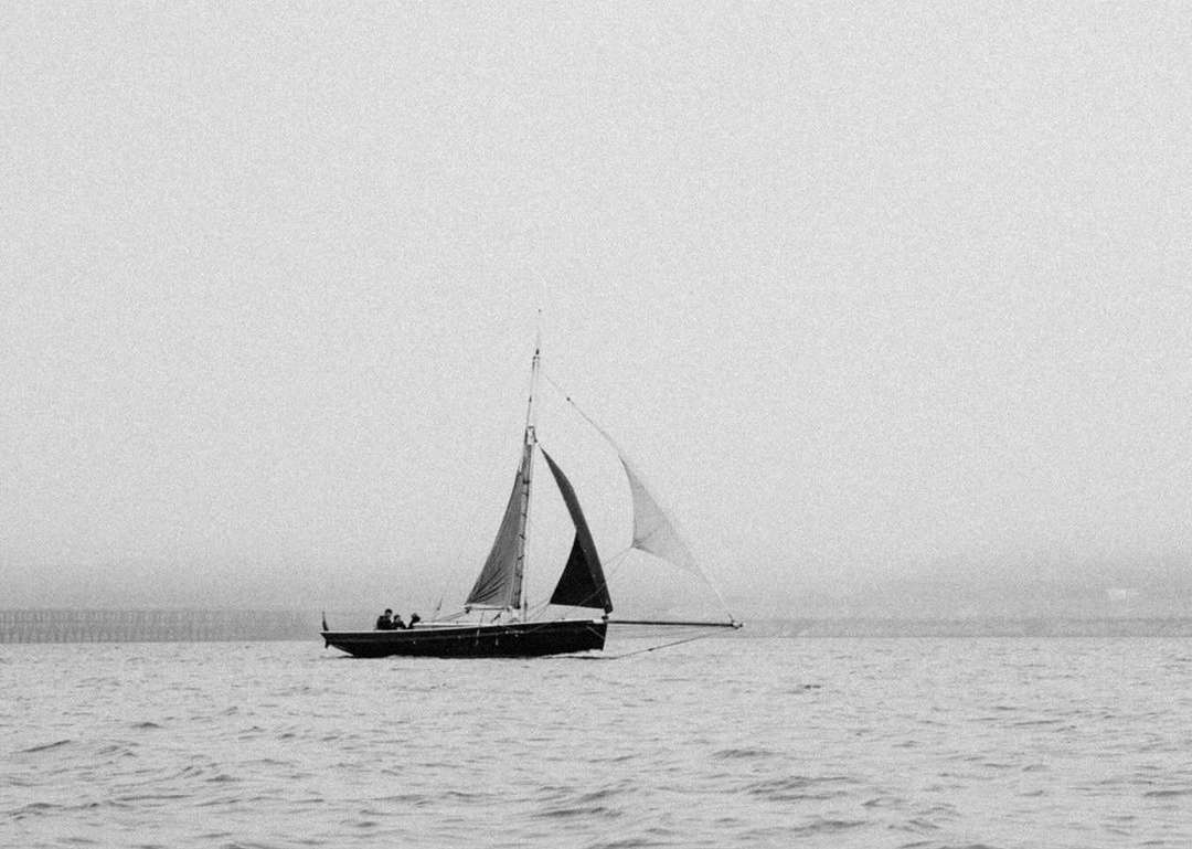 Black and white photograph of a sailboat with two people aboard sailing on the water, with land visible in the distant background.