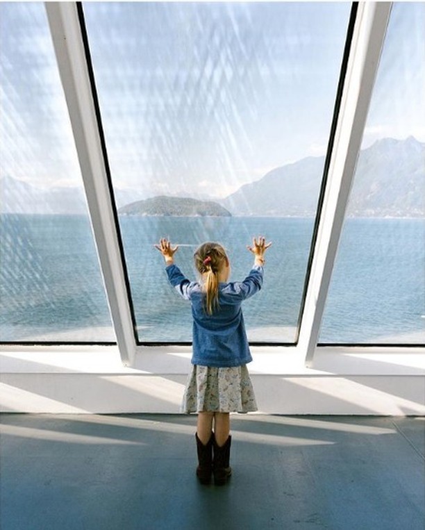 A young girl looks out a window at a lake with mountains in the distance, wearing a denim jacket, patterned dress, and cowboy boots.