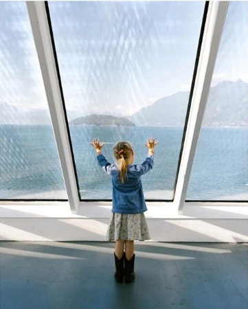 A young girl looks out a window at a lake with mountains in the distance, wearing a denim jacket, patterned dress, and cowboy boots.