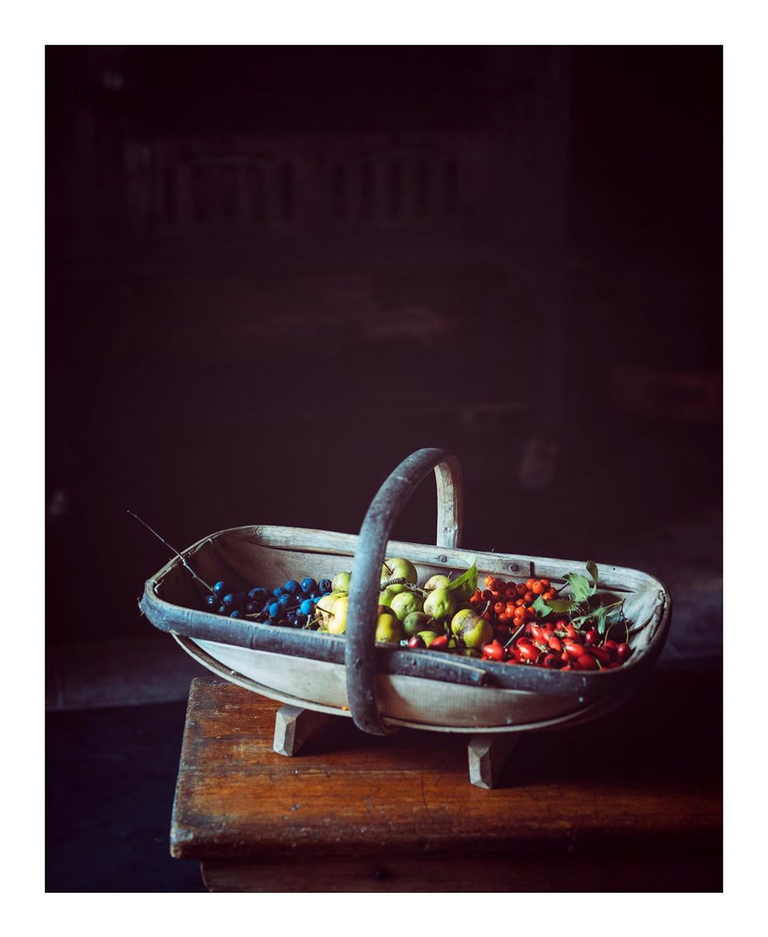 Still life of a rustic basket filled with blueberries, green apples, and red berries on a wooden table in a dimly lit room.