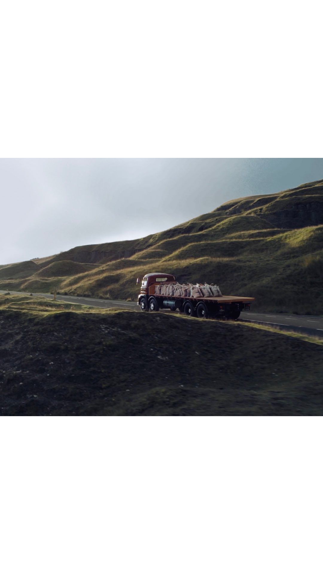 Orange flatbed truck drives on winding road through a green, grassy, hilly landscape. Haze in the air. Nature & Wildlife.
