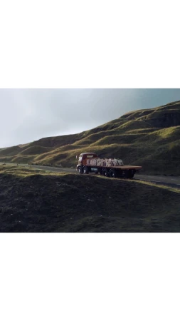 Orange flatbed truck drives on winding road through a green, grassy, hilly landscape. Haze in the air. Nature & Wildlife.