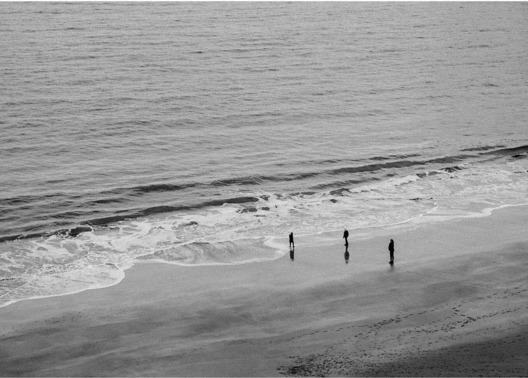Black and white photograph of three people standing on a sandy beach near the ocean, with gentle waves lapping at the shore.