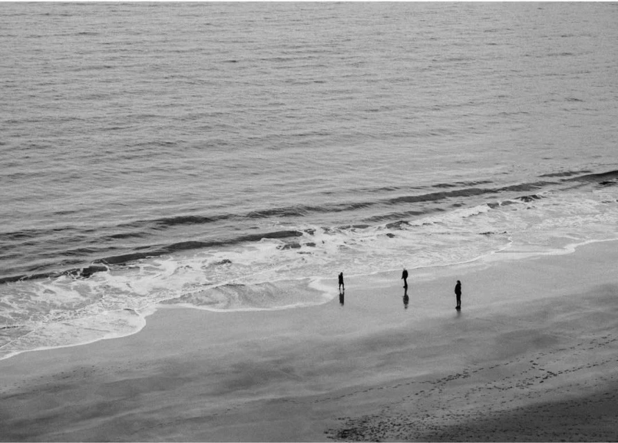 Black and white photograph of three people standing on a sandy beach near the ocean, with gentle waves lapping at the shore.