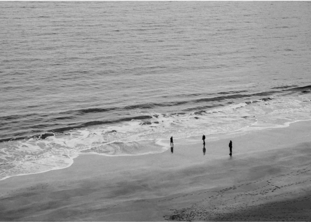 Black and white photograph of three people standing on a sandy beach near the ocean, with gentle waves lapping at the shore.