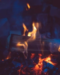 Close-up vertical photograph of a burning log surrounded by bright orange flames and glowing embers against a dark blue background.
