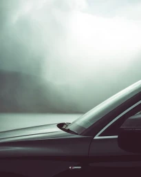 Close-up profile of a dark car parked against a hazy, misty body of water and overcast sky background.