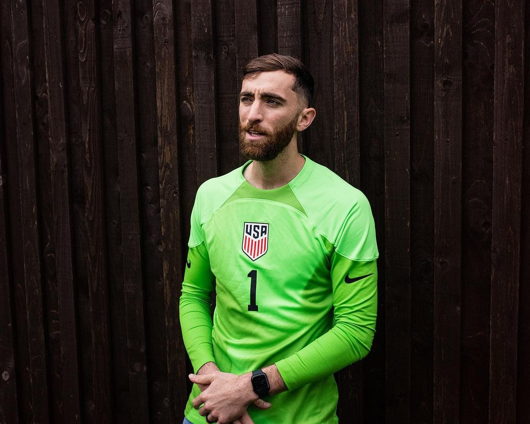 Adult male in athletic gear posing outdoors against dark wooden fence. He is wearing a lime green jersey with the USA logo and number 1.