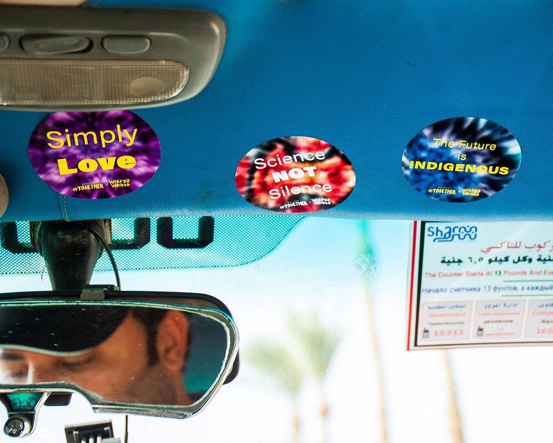 Three stickers with messages of love, science, and indigenous culture, adhered to a car ceiling, with a rearview mirror reflecting a face.