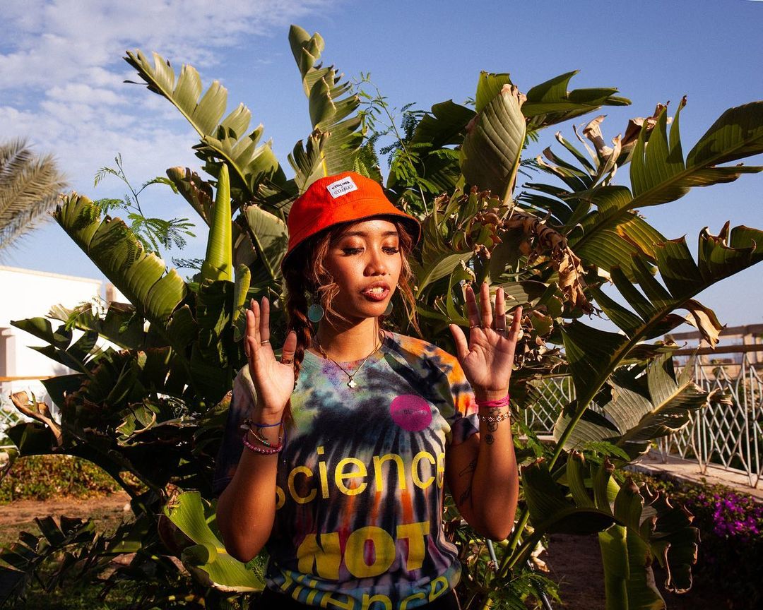 Young woman in an orange bucket hat and tie-dye science t-shirt poses outdoors with palms against a blue sky. Fun, youthful style.