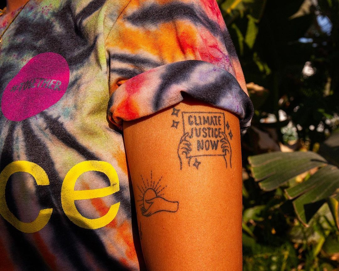 Arm with climate justice tattoo, wearing a tie-dye shirt outside amidst greenery, advocating for environmental action and social responsibility.
