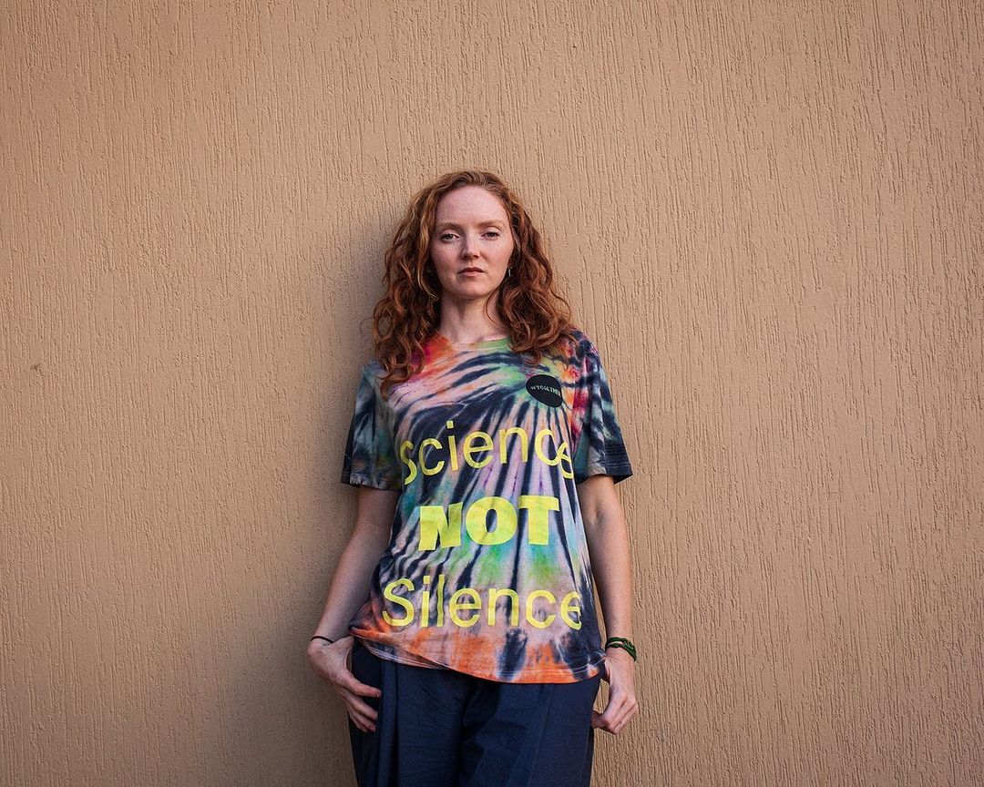 Adult female with red hair wearing a tie-dye T-shirt with the text 'Science Not Silence' standing against a tan wall.