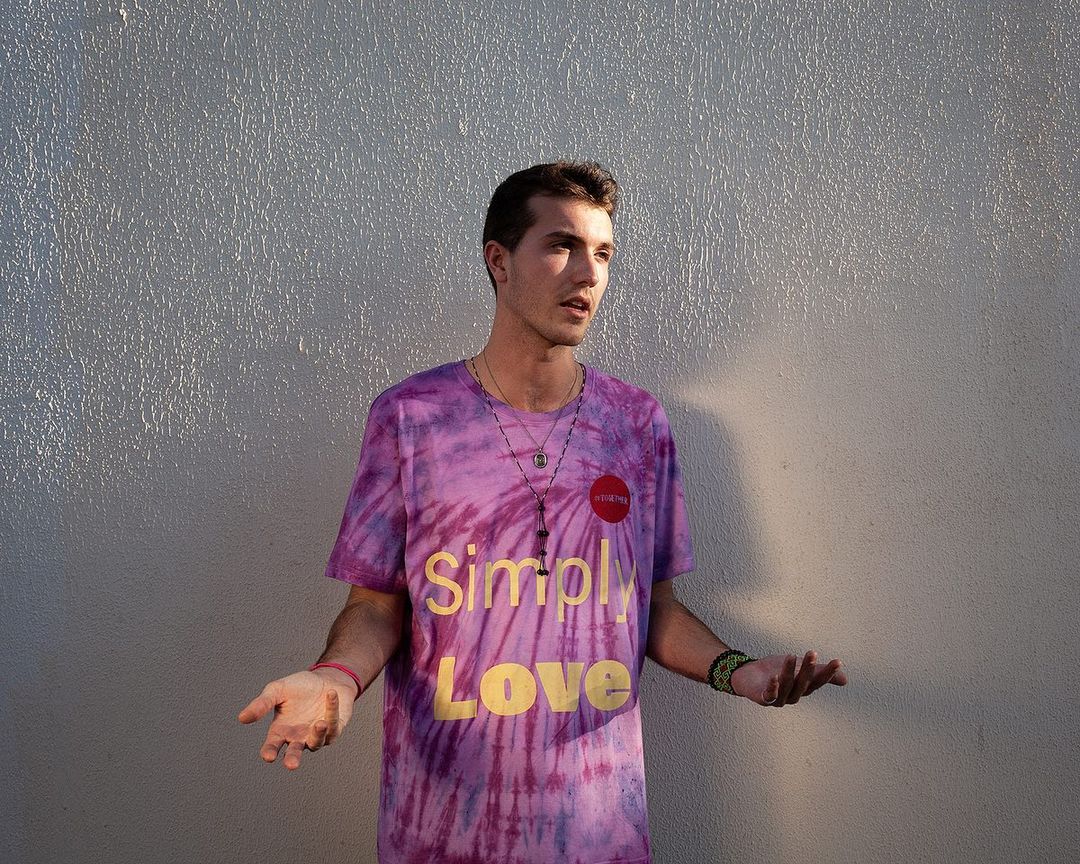 Adult male in a tie-dye shirt with 'Simply Love' text, standing against a textured gray wall, wearing necklaces and a beaded bracelet.