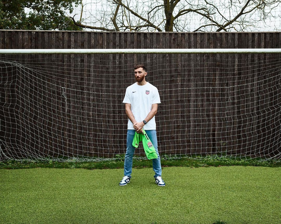 Bearded man in a USA t-shirt stands on a soccer field in front of a net, holding a green jersey, looking away in a portrait shot.