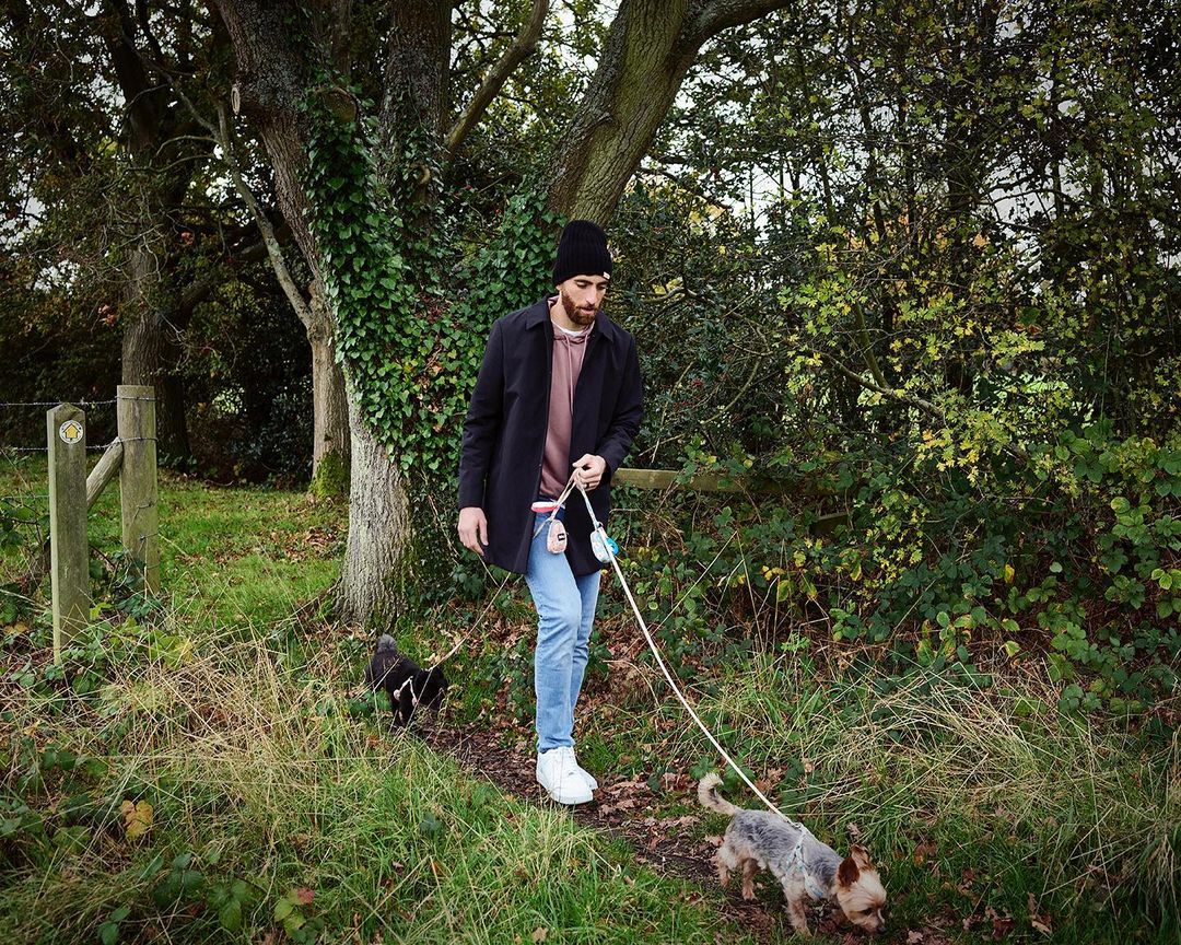 A man in a black coat walks on a path with two small dogs in a leafy, green outdoor setting with a wooden fence.