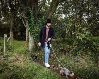 Man wearing a black coat and beanie walking two dogs along a grassy woodland trail