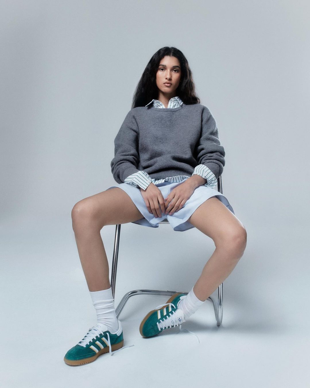 Woman with long dark hair seated on a metal chair wearing a grey sweater over a striped shirt, shorts, and green sneakers.