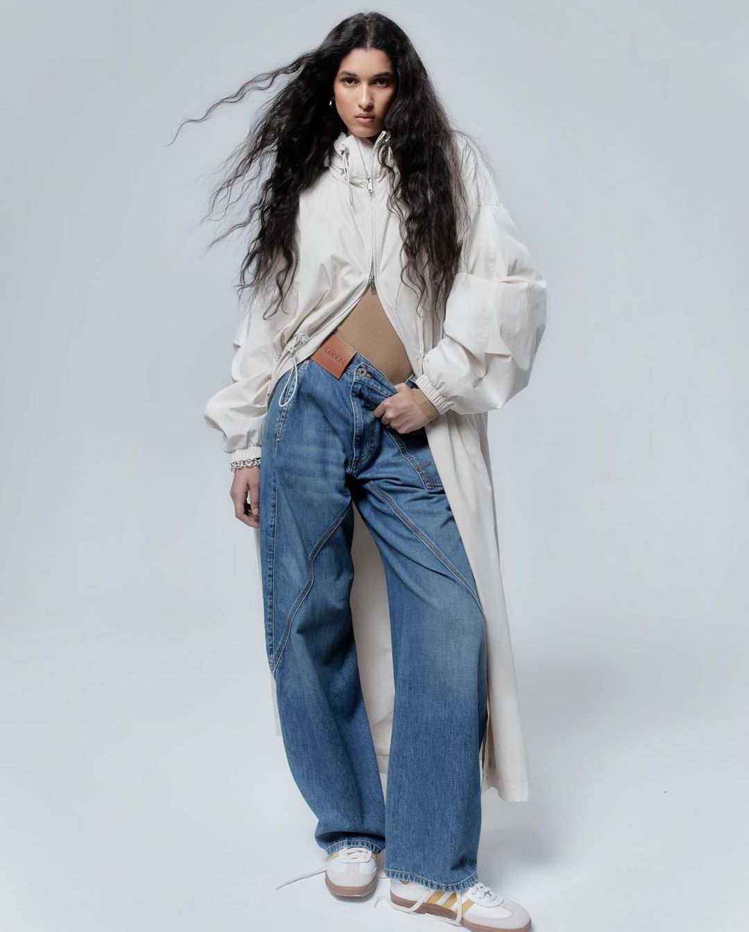 A full shot of a woman posing in baggy jeans and a long white jacket against a bright white backdrop in a fashion editorial style.
