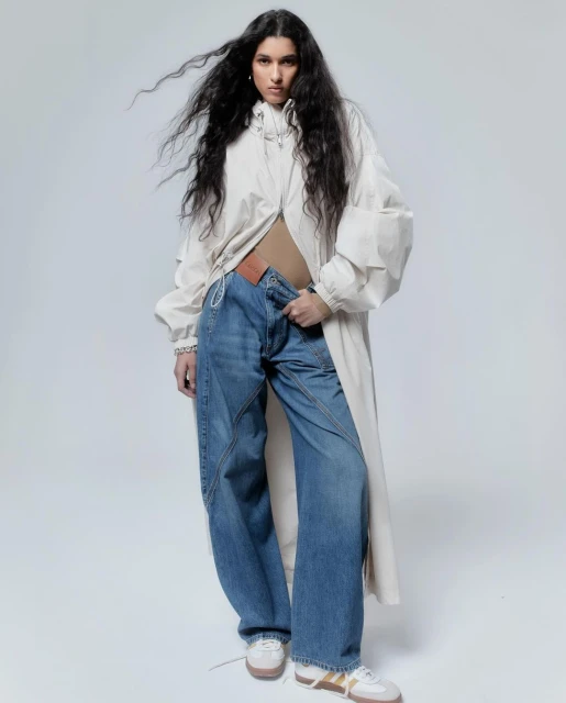 A full shot of a woman posing in baggy jeans and a long white jacket against a bright white backdrop in a fashion editorial style.