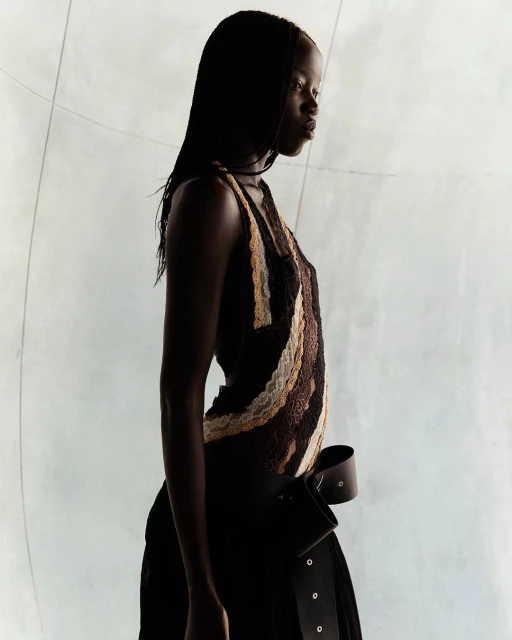 Studio shot of a Black woman in a textured brown and tan dress with a wide leather belt, against a muted, soft-lit neutral background.