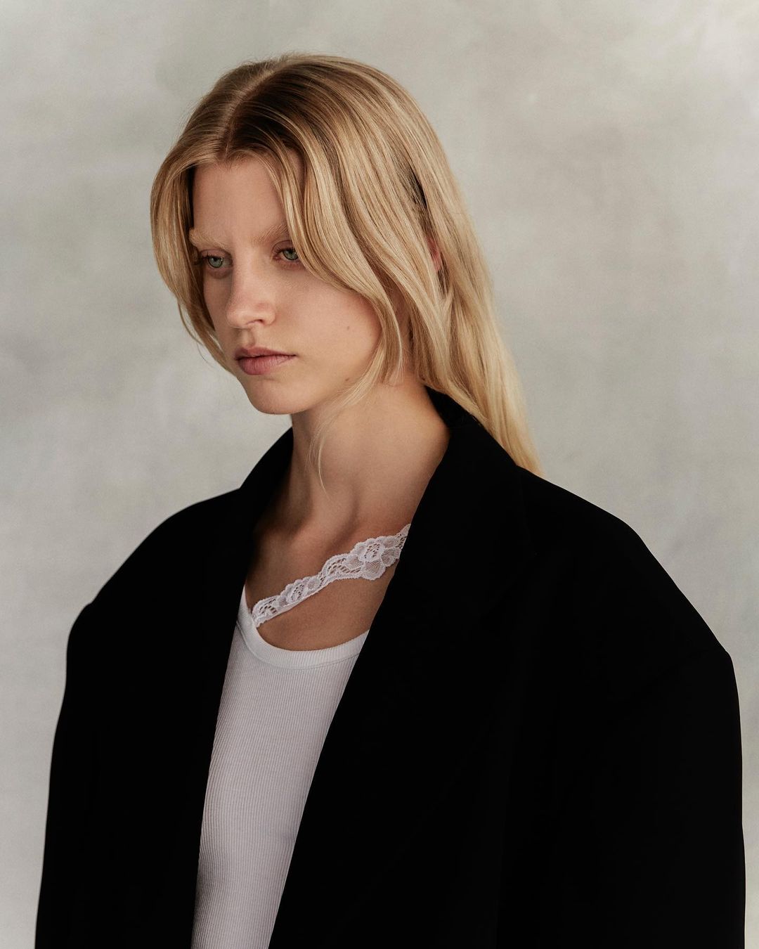 Portrait of a blonde woman wearing a black jacket and white lace-trimmed top against a neutral background.