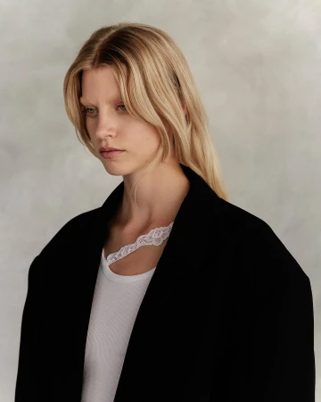 Portrait of a blonde woman wearing a black jacket and white lace-trimmed top against a neutral background.