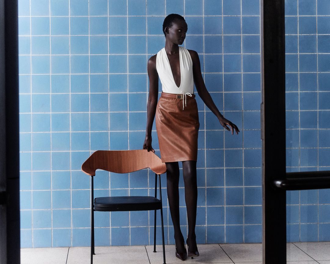 Fashion editorial shot: Black woman poses in a white halter top and tan pencil skirt against a blue tiled wall near a wooden chair.