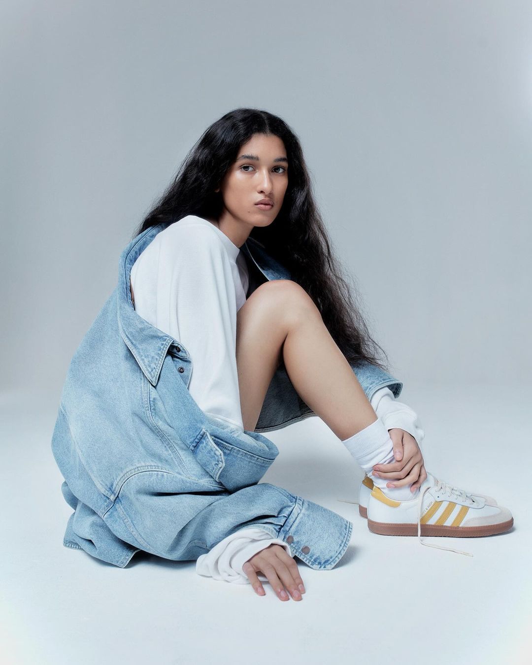 Woman with long black hair sits wearing a white t-shirt, light denim jacket, white socks, and beige and white sneakers in a studio.