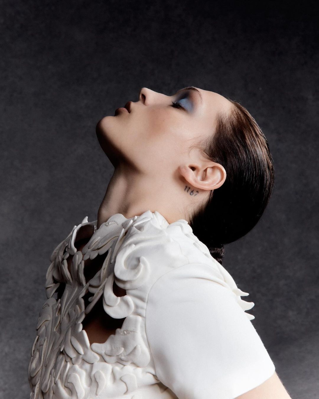 Profile portrait of a woman in a white sculptural top, looking up against a dark grey background in a beauty and fashion context.