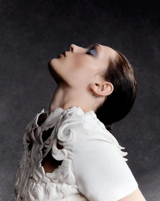 Profile portrait of a woman in a white sculptural top, looking up against a dark grey background in a beauty and fashion context.