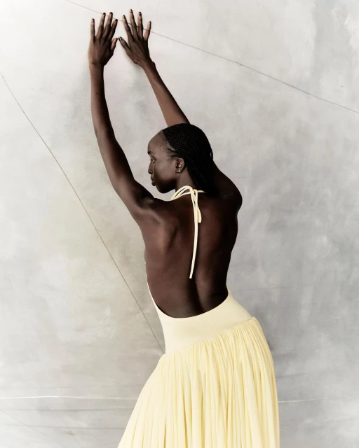 A slender woman with braids in a flowing yellow dress leans against a gray wall with her arms raised above her head in a studio shot.