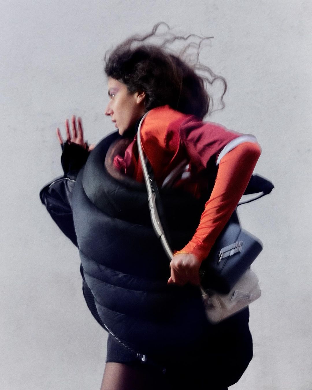 Woman in motion wearing a puffy black vest over a red shirt and black skirt, carrying a white purse with a blurred background.