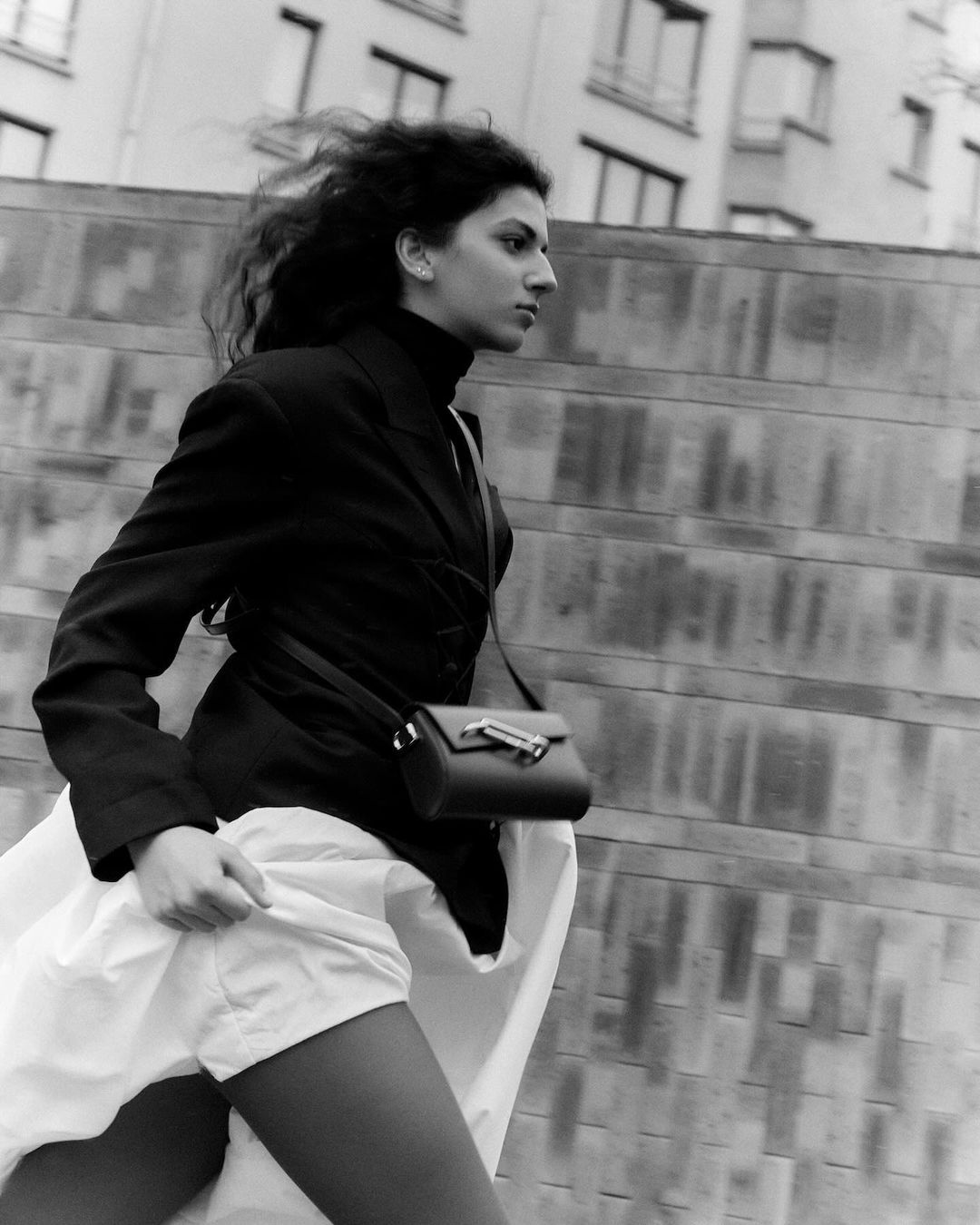 Black and white medium shot of a woman walking outdoors in a blazer, skirt, and crossbody bag, against an urban background.