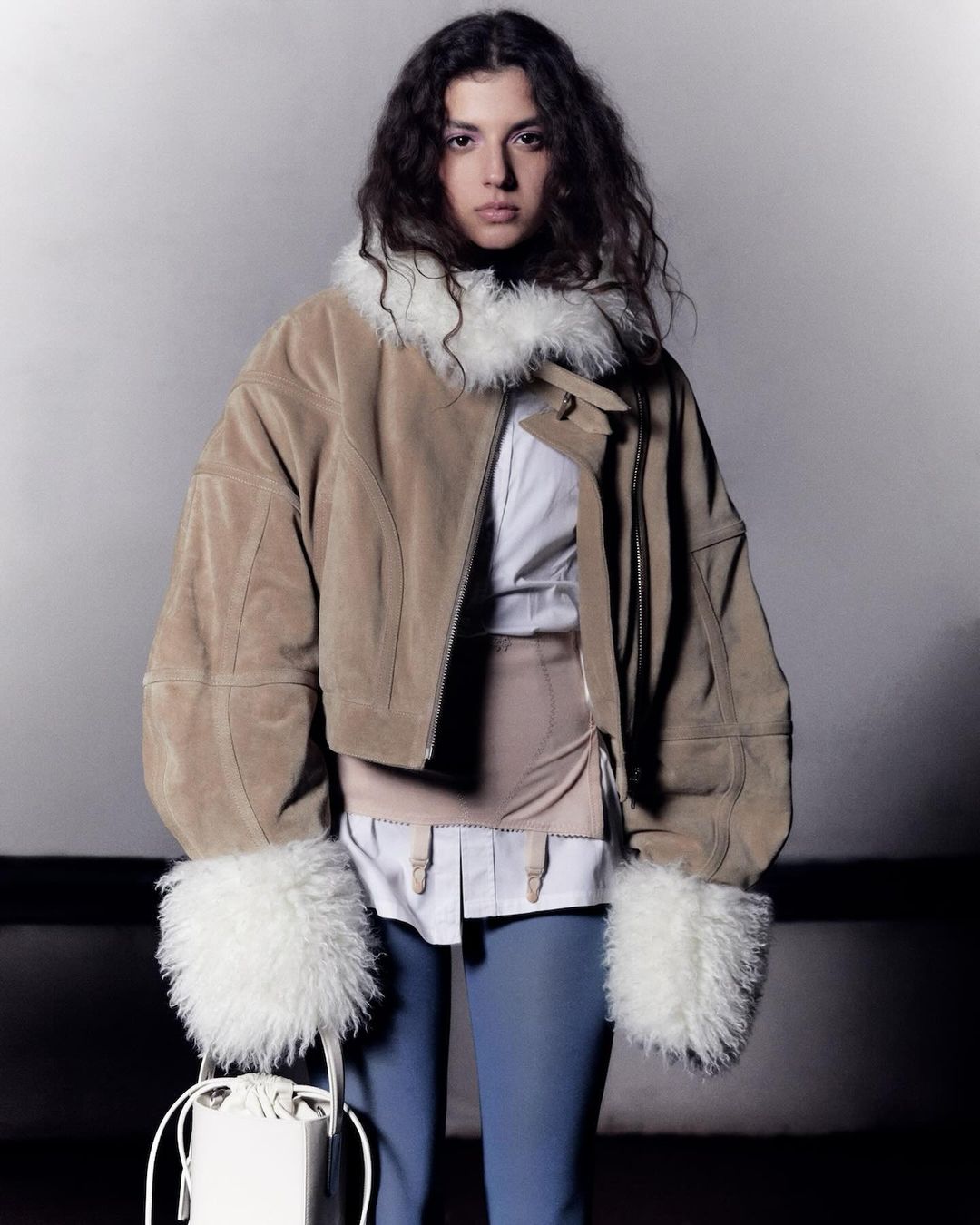 A woman with curly hair poses in a tan suede jacket with white fur trim, paired with light blue pants and carrying a white bag.