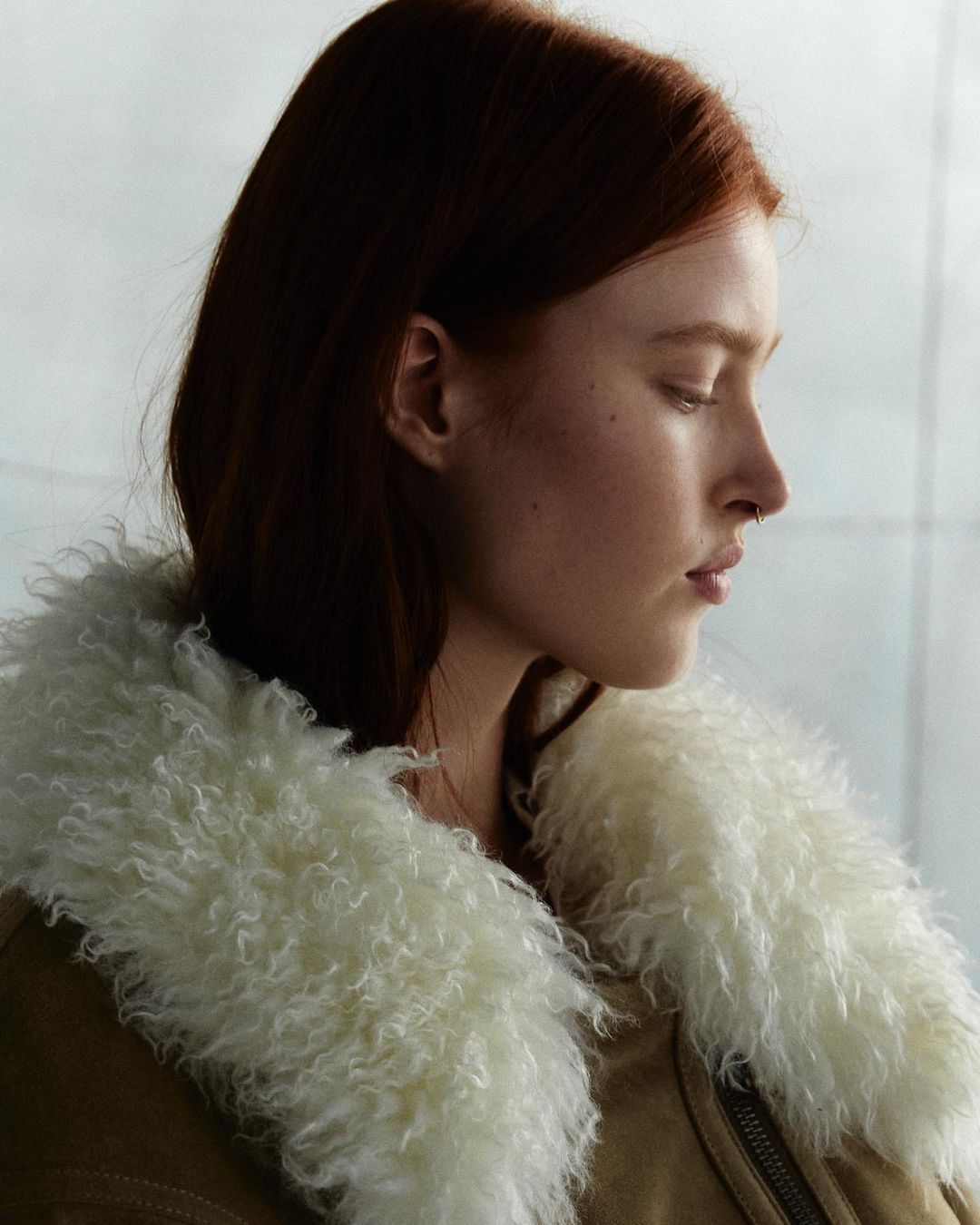 Profile shot of a red-haired woman wearing a fuzzy-collared jacket, looking down against a muted grey-white background with soft lighting.