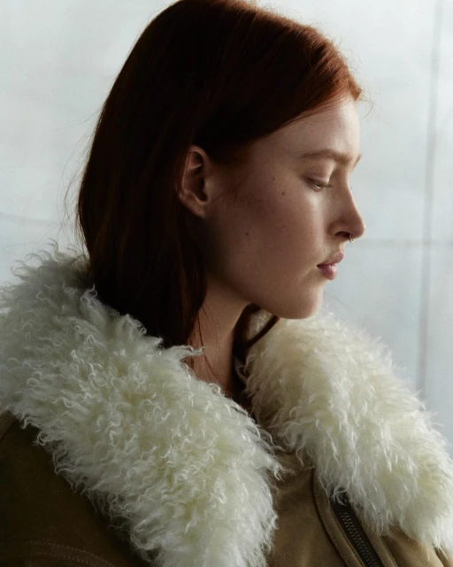 Profile shot of a red-haired woman wearing a fuzzy-collared jacket, looking down against a muted grey-white background with soft lighting.