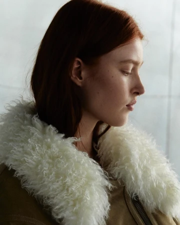Profile shot of a red-haired woman wearing a fuzzy-collared jacket, looking down against a muted grey-white background with soft lighting.