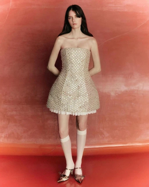 A full shot of a woman posing in a strapless metallic mini dress with white knee socks and embellished heels against a peach-colored background.