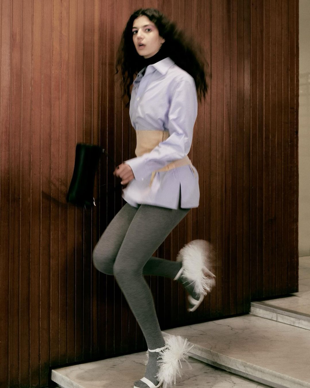 Young woman in fashionable outfit poses indoors, wearing gray tights, white shirt, and white feather-trimmed heels, carrying a black purse.