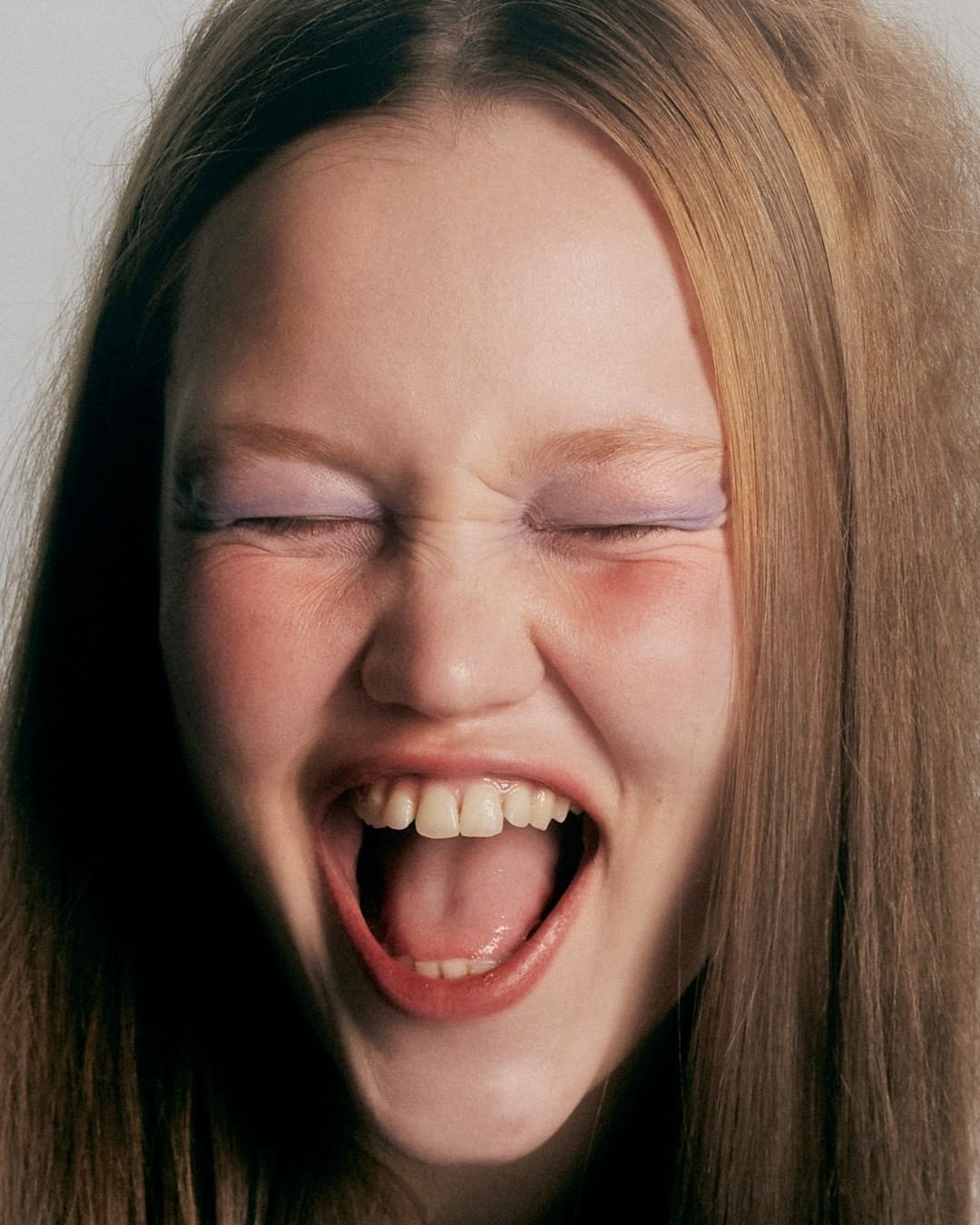 Close-up portrait of a laughing young adult woman with blonde hair, closed eyes, purple eyeshadow, and light skin tone on a white background.
