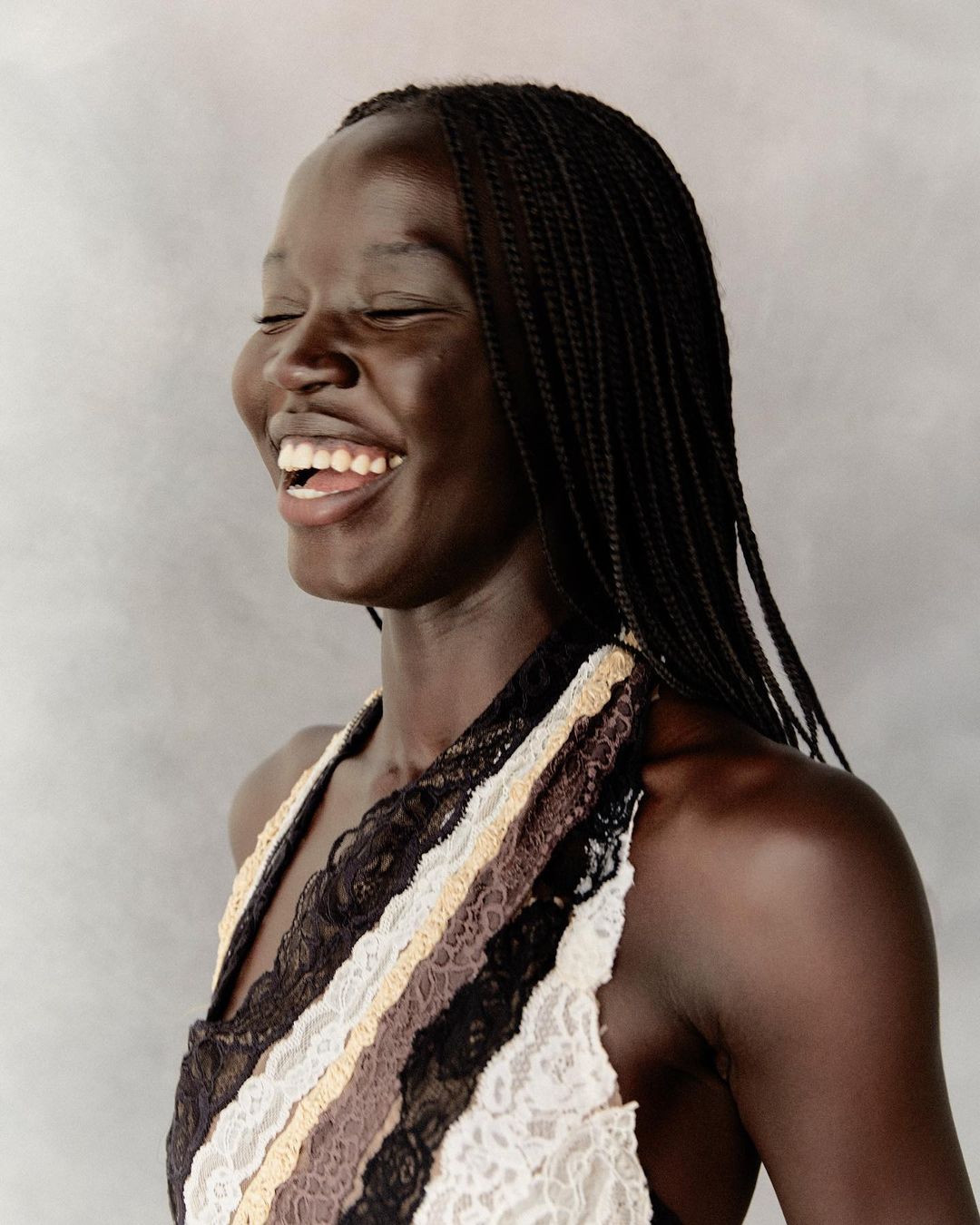 Portrait of a laughing black woman with braided hair wearing a patterned lace halter top against a textured grey background.