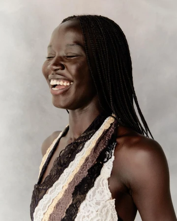 Portrait of a laughing black woman with braided hair wearing a patterned lace halter top against a textured grey background.