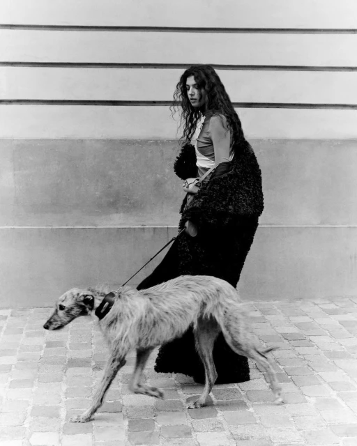 Monochrome shot of a woman walking with a dog on a leash, wearing a fur coat, on a brick sidewalk with a wall in the background.