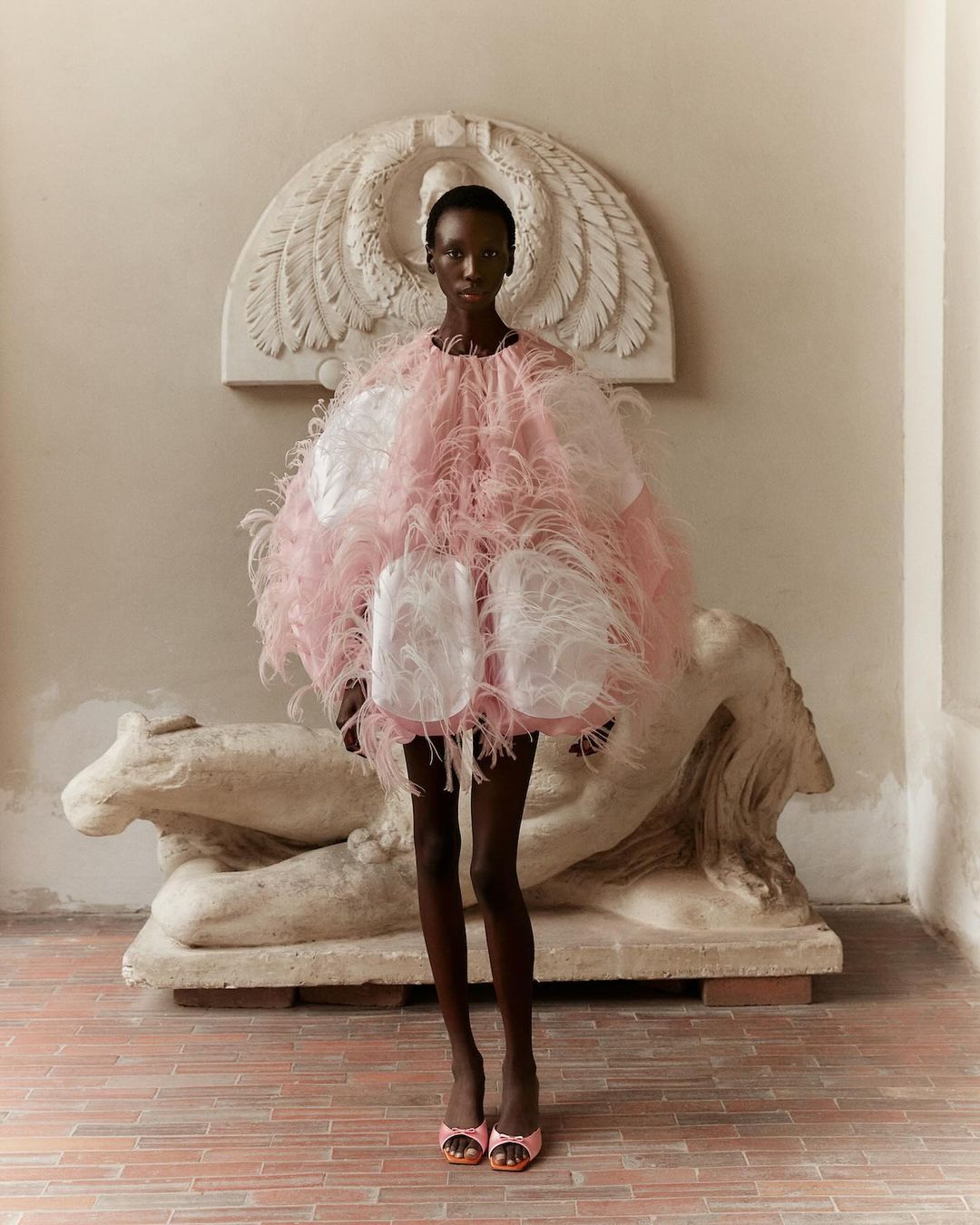 A woman in a feathered pink and white dress poses in front of an antique stone sculpture in an elegant interior setting.