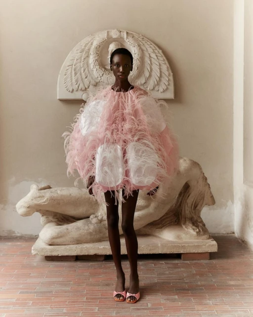 A woman in a feathered pink and white dress poses in front of an antique stone sculpture in an elegant interior setting.