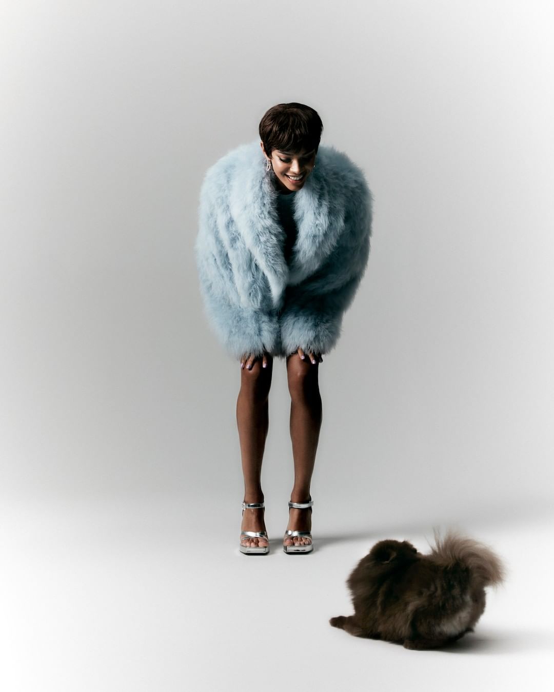 A fashionable adult woman in a light-blue fur coat smiles down at a fluffy brown dog on a white surface in a studio setting.
