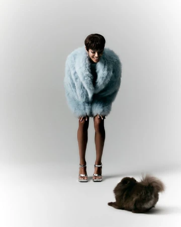 A fashionable adult woman in a light-blue fur coat smiles down at a fluffy brown dog on a white surface in a studio setting.