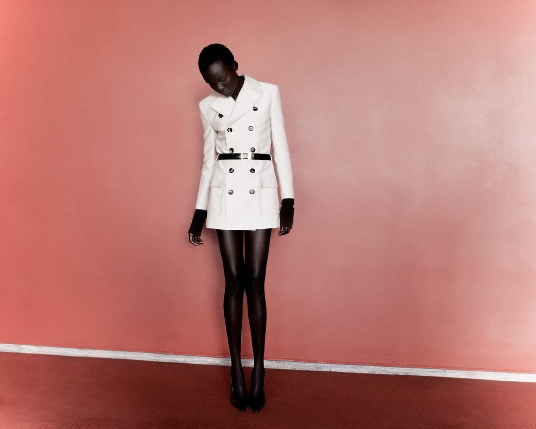 A slender Black woman in a white double-breasted dress stands in front of a peach wall, captured in a full shot, showcasing elegance.