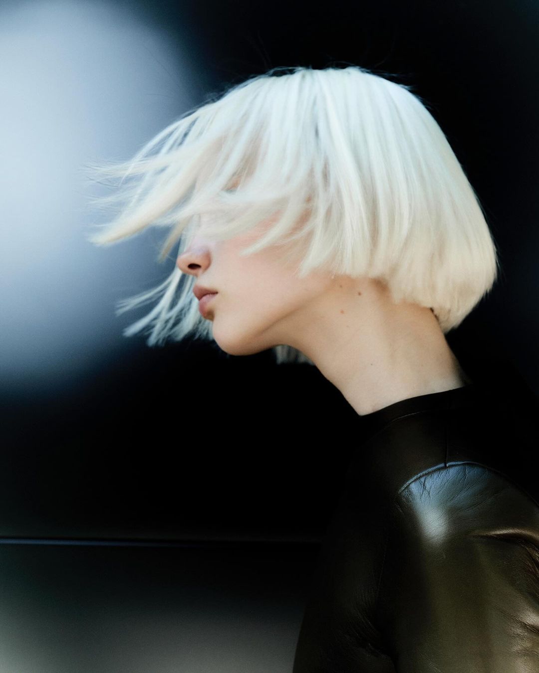 Profile of a woman with short platinum blonde hair blowing, wearing a black top against a dark background with a blurred light.