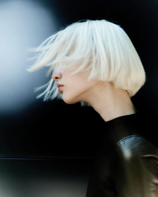 Profile of a woman with short platinum blonde hair blowing, wearing a black top against a dark background with a blurred light.